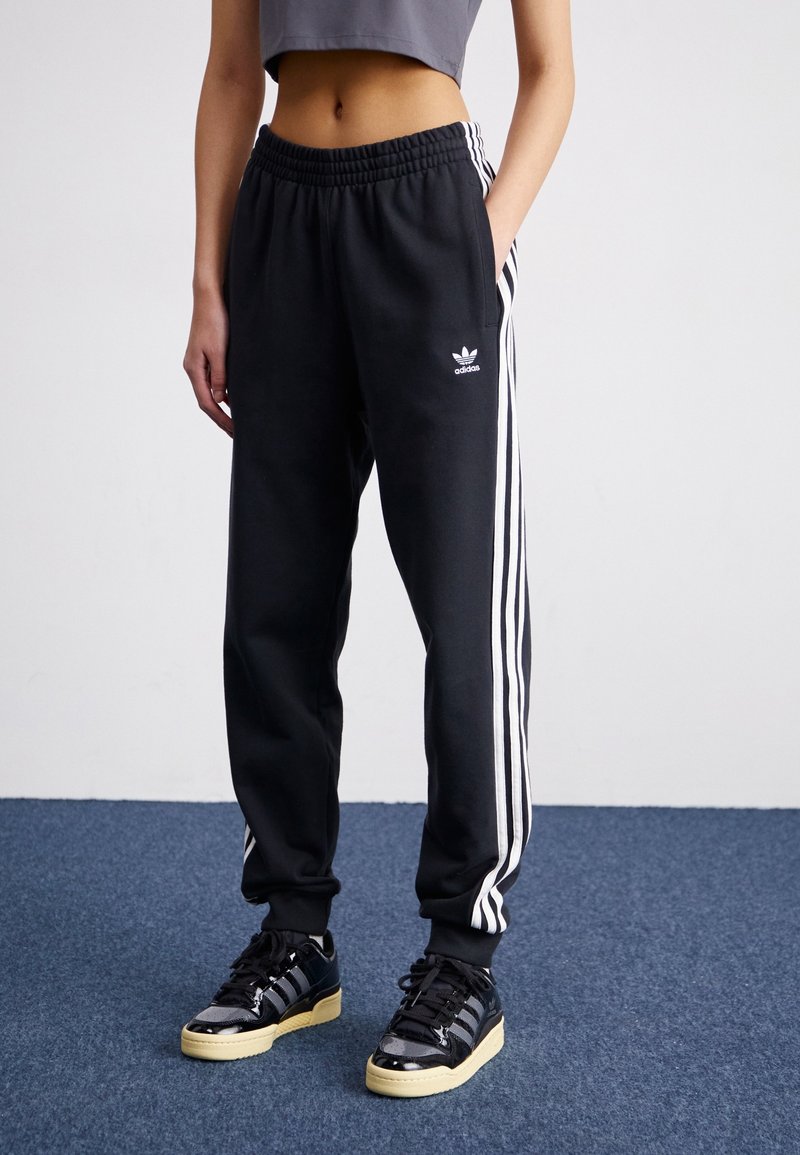 Svarta sweatpants med vita sidostripes, elastisk midja och fickor. Kombinerad med svarta sneakers med glänsande yta och gummisålar.