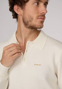 Polo en tricot de couleur crème avec un col classique, un tissu texturé et trois petites broderies d'animaux marron sur la poitrine.