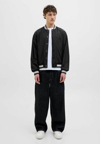 Veste bomber noire avec des rayures blanches sur le col et les poignets, portée sur un t-shirt blanc ; associée à un pantalon en denim noir ample et des baskets blanches.