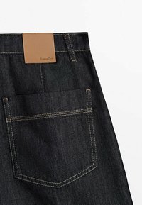 Jean en denim foncé avec une finition texturée, comportant une pièce en similicuir beige au niveau de la taille et une poche arrière unique avec des coutures contrastées.