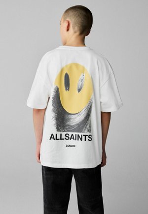 Mladá osoba s krátkymi vlasmi nosiaca biele tričko AllSaints s veľkým žltým usmievavým tvárou a čiernou grafikou na chrbte.
