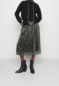 Suéter negro de punto, falda midi plisada metálica en verde oscuro, botas negras de tobillo con punta afilada y un pequeño bolso cruzado negro.