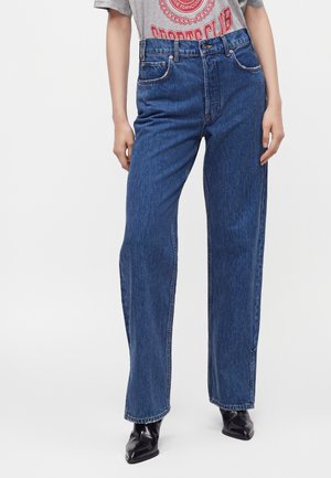 Personne portant un jean bleu taille haute à jambes droites avec un t-shirt gris de club de sport rentré, associé à des bottes noires à bout pointu.