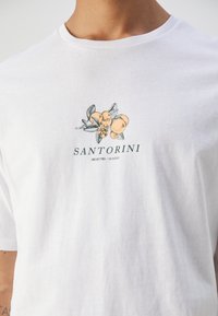 Bílý bavlněný tričko s texturovaným botanickým potiskem v oranžové a zelené barvě. Text "SANTORINI" je vytištěn pod designem.