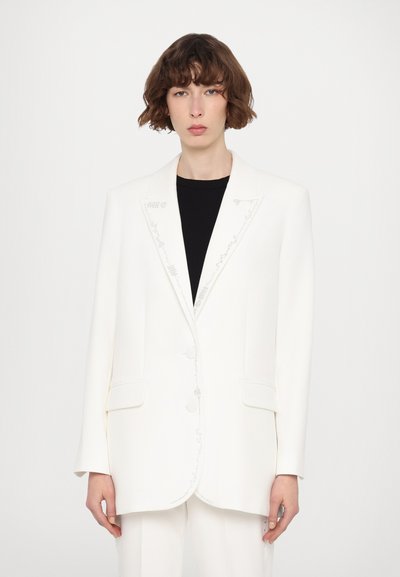 Blazer bianco su misura con un design strutturato, caratterizzato da ricami argentati lungo i rever, due tasche frontali e grandi bottoni.