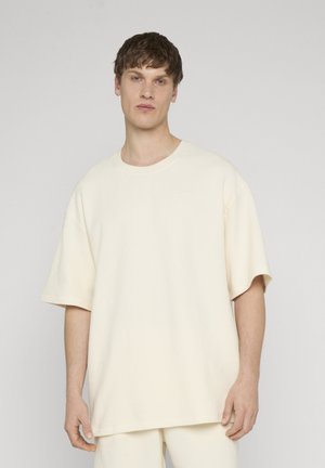 Młody mężczyzna o krótkich brązowych włosach, ubrany w za dużą kremową prążkowaną koszulkę z krótkim rękawem i pasujące spodnie, stojący na jednolitym tle.