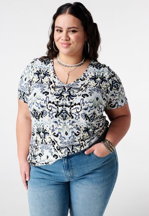 Lachende vrouw met lang donker haar draagt een kort mouwtop met patroon, blauwe spijkerbroek, oorringen, gelaagde kettingen en armbanden, hand in de zak.