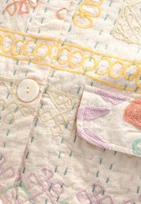 Bestickter Stoff in Hellbeige mit mehrfarbigen Mustern, versehen mit einem weißen Knopf und einer Tasche, die durch pastellfarbene Stickereien betont wird.