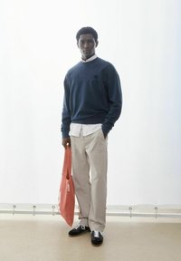 Marineblauwe sweatshirt met logo, wit overhemd met kraag daaronder, beige cargobroek en zwarte sneakers met witte veters, met een koraalkleurige draagtas in de hand.