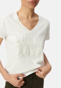 Camiseta blanca de algodón con cuello en V y mangas cortas, que presenta un texto elevado y bordado "LUCKY LOVE" en un estilo texturizado en la parte frontal.