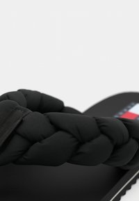 Svart flip-flop med en flätad, vadderad rem och en solid svart sula med en textil logotypaccent i röd och blå.