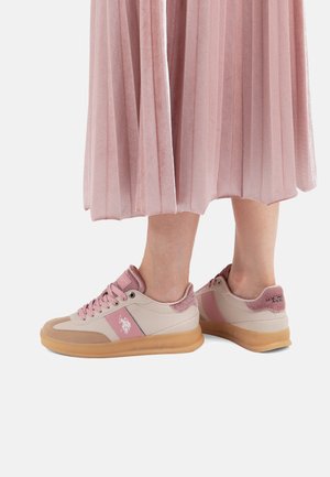 Persona che indossa sneakers rosa e beige con suole in gomma e una gonna plissettata rosa visibile dalla parte inferiore delle gambe.