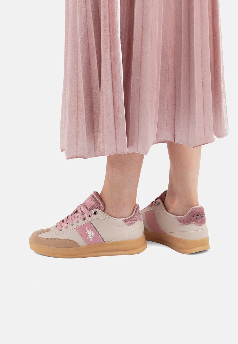 Person, die pinke und beige Sneakers mit Gummisohlen sowie einen pinken, plissierten Rock trägt, der bis zu den Waden sichtbar ist.