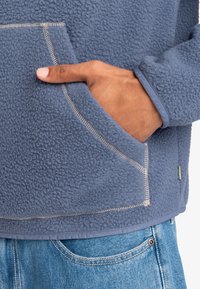 Polarowy pulower w stonowanym niebieskim kolorze z kieszenią z przodu, kontrastowym beżowym przeszyciem i prążkowanymi mankietami, w zestawie z jasnymi dżinsami.