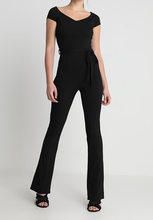 Kobieta ubrana w dopasowany, czarny kombinezon z krótkim rękawem, wiązany w talii, z rozszerzanymi nogawkami, w połączeniu z czarnymi sandałami na obcasie z odkrytymi palcami.