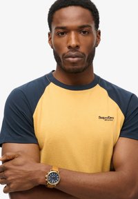 Camiseta de manga corta con cuerpo amarillo y mangas raglán en azul marino. Logo bordado en el pecho. Reloj dorado con correa de cuero en la muñeca.