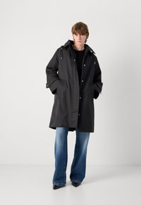 Black waterproof coat with a hood, featuring snap buttons and side pockets. Worn over a black top and paired with wide-leg jeans.