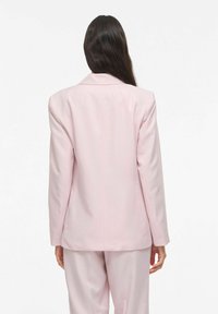 Blazer rose clair sur mesure avec épaules structurées, fermeture à un bouton et finition en tissu lisse. La vue de dos révèle des lignes épurées sans ornements.