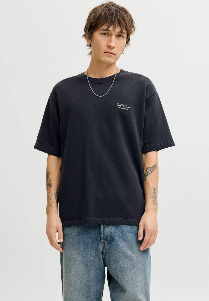 Jeune homme aux cheveux bruns courts portant un t-shirt noir Jack & Jones, une chaîne argentée, des boucles d'oreilles et un jean bleu, debout devant un fond uni.