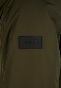 Tissu vert olive avec une pièce rectangulaire noire cousue dessus, affichant le nom de la marque "HECHTER" en lettres en relief.
