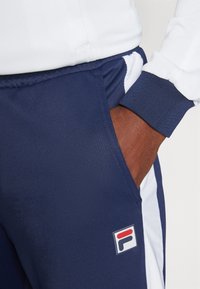 Navy track pants with white side stripes, made of smooth fabric. Features pocket and a multicolored FILA logo on the left thigh.