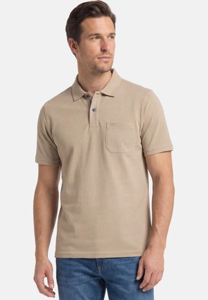 Mand iført beige poloshirt med korte ærmer og brystlomme samt blå jeans, kigger let mod højre, neutral baggrund.