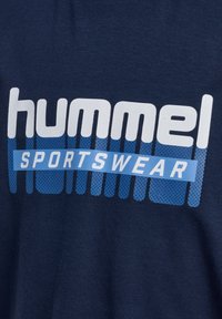 Dunkelblaues Sportoberteil mit weißem und blauem Text "hummel SPORTSWEAR", das ein Punktemuster und ein rechteckiges Designelement aufweist.