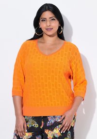 Orange V-Ausschnitt-Pullover mit strukturiertem Muster, Dreiviertelärmeln und geripptem Saum. Kombiniert mit bunten Hosen mit Schmetterlingsdruck.