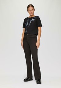Schwarzes T-Shirt mit kurzen Ärmeln, blauer Schrift und Verzierungen, kombiniert mit dunkelbraunen Schlaghosen und schwarzen klobigen Schuhen.