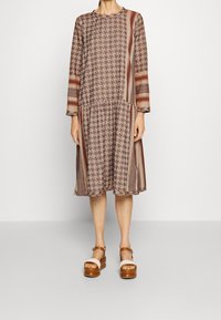 Hnědé šaty s vzorem a dlouhými rukávy, které kombinují kostkovaný a pruhovaný vzor. Tkanina má lehký průběh, doplněná béžovými platformovými sandály.