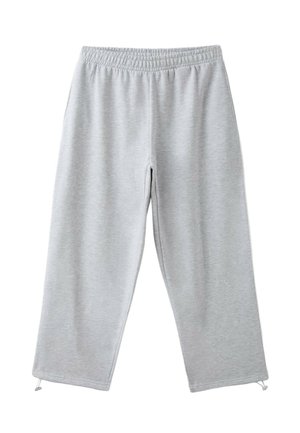 Personne portant un haut à manches longues olive et un t-shirt court blanc avec un pantalon de jogging gris clair, main partiellement glissée sous le t-shirt près de la taille.