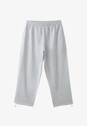 Personne portant un haut à manches longues olive et un t-shirt court blanc avec un pantalon de jogging gris clair, main partiellement glissée sous le t-shirt près de la taille.