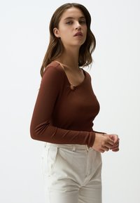 Braun gestreiftes Langarmoberteil mit rundem Halsausschnitt und dekorativem Verschluss an der Schulter, kombiniert mit hochtaillierten weißen Jeans.