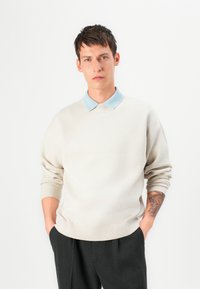 Pullover bege com uma camisa azul de colarinho por baixo, apresentando uma textura suave e um corte relaxado, usado com calças escuros de corte clássico.