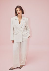 Dubultā pārsega gaiši bežs bleizers un atbilstošas plašas bikses, kas izgatavotas no mīkstas auduma; rotāts ar zelta toņa pogām un kabatām. Akcentēts ar čūskas drukas apaviem.