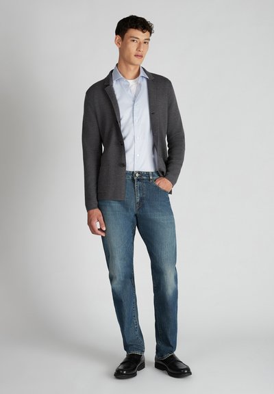 Grauer Cardigan mit zwei Knöpfen, hellblauem Hemd, blauer Jeans und schwarzen Schuhen. Weicher Stoff, lässige Passform und kontrastierende Farben.