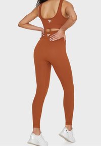 Brązowy zestaw sportowy z prążkowaniem, składający się z biustonosza i legginsów o dopasowanym kroju. Widoczne akcenty białego logo na plecach. Noszony z białymi butami sportowymi.