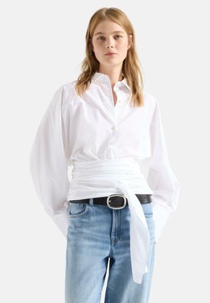 Jonge vrouw die een witte blouse met knopen, pofmouwen en een omslagriem draagt, gecombineerd met lichtblauwe jeans en een zwarte riem.