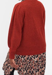 Czerwony sweter z dzianiny z bufiastymi rękawami, prążkowanymi mankietami i dolnym brzegiem. Zestawiony z czarną spódnicą w beżowy i różowy abstrakcyjny wzór w panterkę.