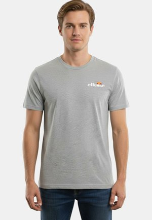 Uomo con una t-shirt ellesse grigio chiaro e jeans blu scuro in piedi su uno sfondo bianco semplice.