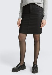 Jupe crayon en denim noir avec poches avant et finition lisse. Associée à des mocassins noirs et des collants noirs transparents.