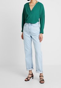 Person in einem grünen Langarmblusendesign, mit hellblauen hochgekrempelten Jeans, schwarzen Riemchen-Heels und einer goldenen Kettenhalskette vor einem weißen Hintergrund.