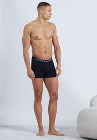 Μυώδης άνδρας όρθιος, ξυπόλυτος, φορώντας μαύρα αντρικά εσώρουχα Tommy Hilfiger, κοιτάζει αριστερά του μπροστά σε ένα απλό φωτεινό φόντο.