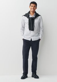 Chemise à rayures blanches et grises à boutons, pull gris foncé posé sur les épaules, pantalon bleu marine et chaussures noires à lacets. Fond neutre.