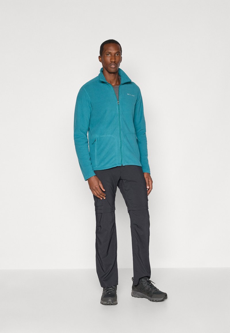 Turkusowa polarowa kurtka z zamkiem, z dwoma przednimi kieszeniami, zestawiona z czarnymi spodniami z zamkiem błyskawicznym i czarnymi butami turystycznymi. Gładka tekstura, swobodny krój.