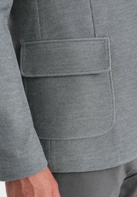 Grau beschichtete Jacke mit strukturiertem Muster, einer seitlichen Pattentasche und tailliertem Design. Der Ärmel weist einen sauberen Abschluss auf.