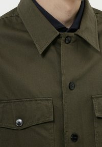 Camisa verde oliva con cuello puntiagudo, botones negros, dos bolsillos en el pecho y detalles de costura visibles. El material parece resistente y texturado.