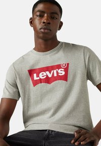 Joven vestido con una camiseta gris de Levi's con un logo rojo y blanco, sentado con una mano apoyada en su muslo contra un fondo liso.