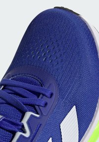 Blauer Sportschuh mit strukturiertem Strick-Obermaterial, sichtbaren Perforationen, lila Schnürsenkeln und einem kontrastierenden weißen Logo an der Seite.
