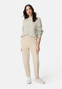 Femme aux longs cheveux foncés portant un pull en maille gris clair, un pantalon cargo beige et des baskets blanches, debout devant un fond blanc uni.